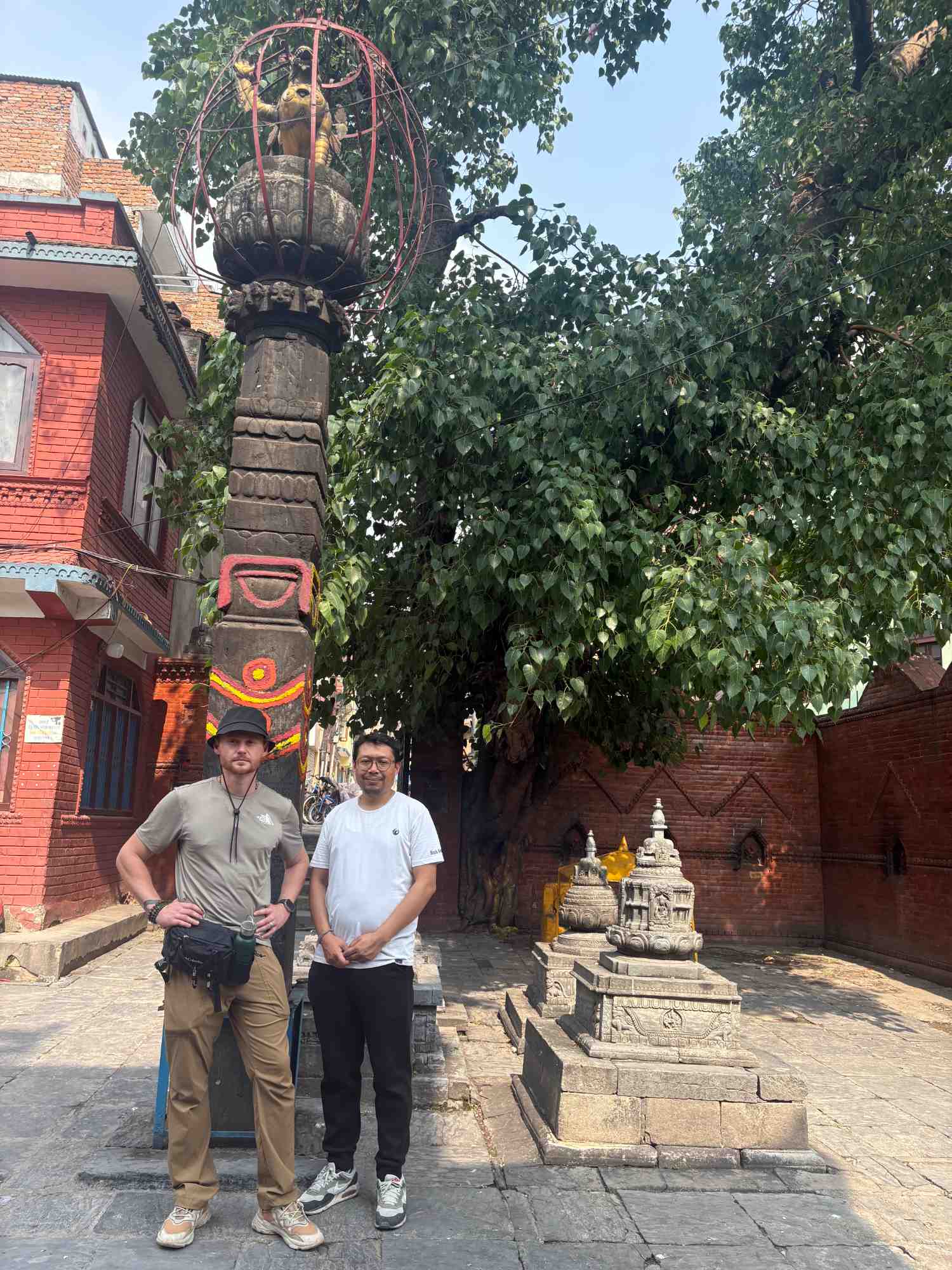 Two men standing beside a carved stone pillar and votive caityas at the Kankesvari temple in Kathmandu