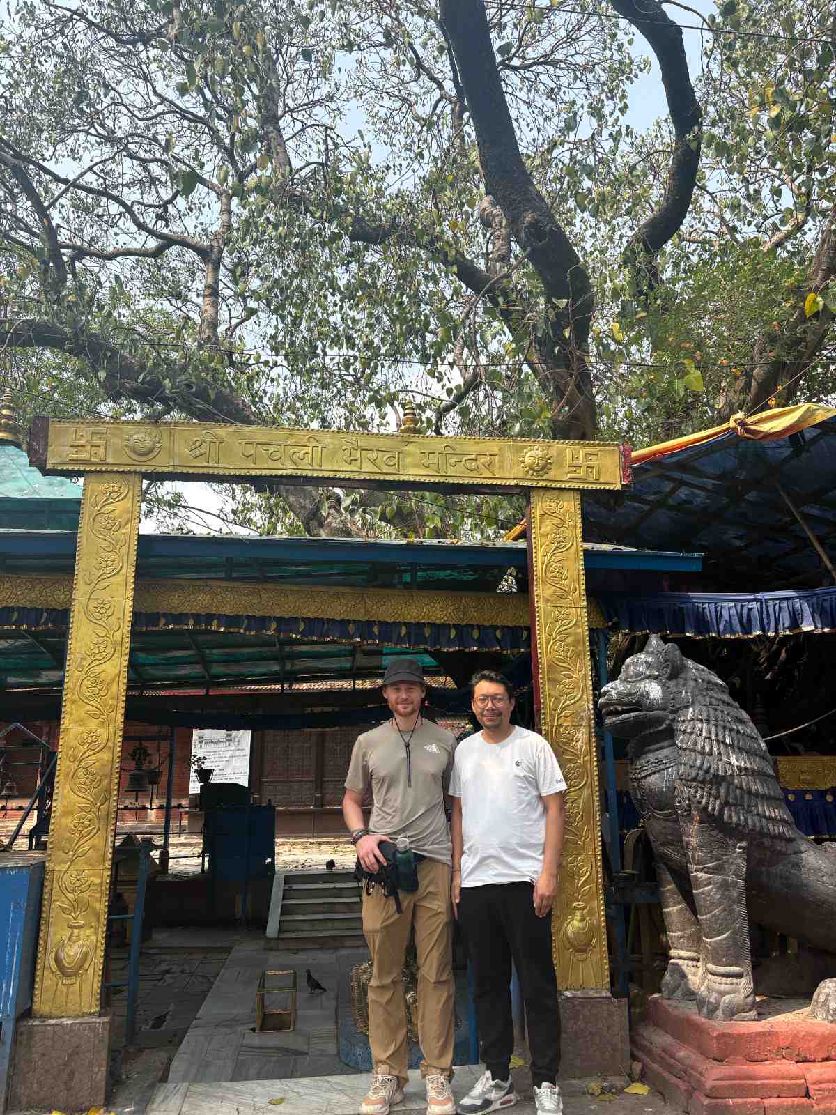 Pilgrims standing at the gilded gateway of the Pachali Bhairava temple in Kathmandu, beneath the old pipal tree